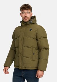 Mand iført en olivengrøn dunjakke med hætte, sort logo-patch på brystet og blå jeans, stående imod en ensfarvet baggrund.