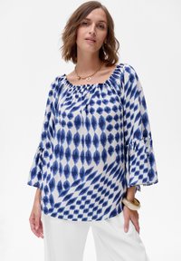 Biała bluzka z niebieskim geometrycznym wzorem, z dekoltem w serek i rękawami typu dzwon. Lekki materiał o gładkiej fakturze. Do zestawu dołączone srebrne akcesoria.
