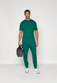 Grønt treningsantrekk som består av en kortermet t-skjorte og matchende joggebukser, laget av et glatt og elastisk materiale. Svart logodetalj.