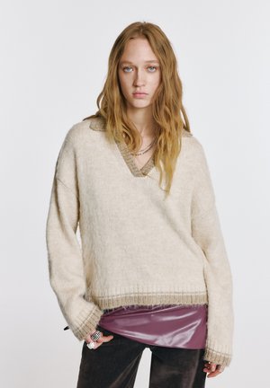 Oversized crème trui met een V-hals en gestructureerde brei, gelaagd over een glanzende bordeauxrode top. De mouwen zijn licht verlaagd met geribbelde boorden.