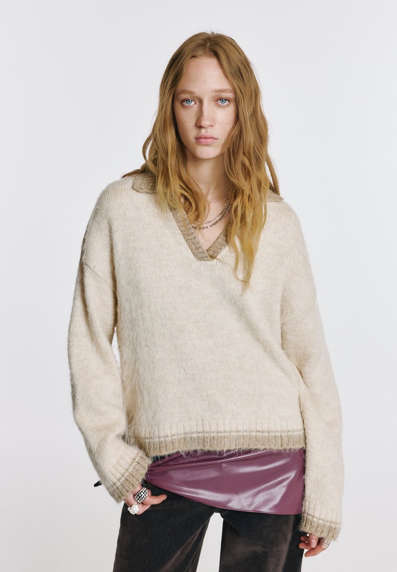 Nagy méretű krémszínű pulóver V-nyakkivágással és texturált kötött mintával, fényes bordó felső rétegeként viselve. Az ujjai enyhén lefelé eresztettek, bordázott mandzsettával.