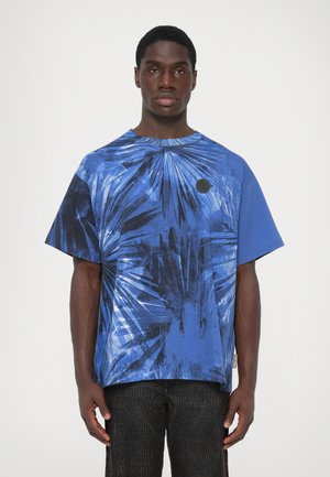 Homem vestido com uma camisa de manga curta azul com um padrão de folhas de palmeira pretas, de pé contra um fundo claro e liso.