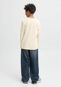 Pojke med kort lockigt hår som bär en krämfärgad långärmad skjorta, lösa mörkblå jeans och vita sneakers, står vänd bort från kameran mot en enkel bakgrund.
