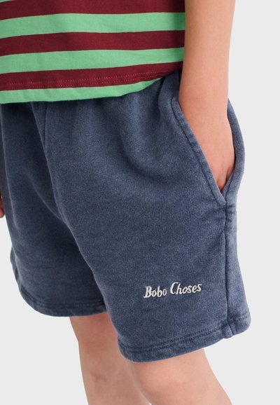 Enfant portant un short bleu avec le logo "Bobo Choses" sur la jambe et une chemise rayée verte et rouge, main placée dans la poche du short.