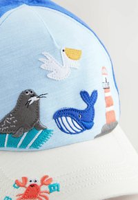 Lyseblå kasket med broderede havdyr-designs: en sæl, hval, pelikan, krabbe og et fyrtårn, med en cremefarvet skygge.