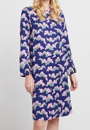 Kvinde iført knælange, langærmede kjole med blå, pink og lysegrå abstrakt skalleret mønster.