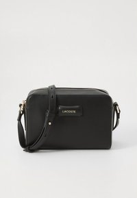 Geantă crossbody din piele neagră, cu fermoar auriu, curea reglabilă și formă dreptunghiulară. Prezintă un accent cu logo-ul Lacoste.