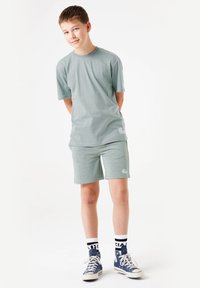 Camiseta de algodón verde claro y pantalones cortos a juego, con un detalle de logo. Zapatillas blancas con acentos azules y calcetines de rayas negras completan el look.
