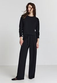 Donna con capelli scuri e sciolti, che indossa una maglietta nera a maniche lunghe, pantaloni neri larghi con cordoncino e scarpe nere basse su uno sfondo semplice.