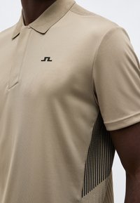 Μπεζ κοντομάνικο πόλο μπλουζάκι με μικρό μαύρο λογότυπο στο στήθος και μαύρη ρίγα κάτω από το μπράτσο.