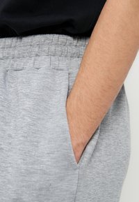 Το χέρι ενός ατόμου ακουμπά στην πλαϊνή τσέπη ανοιχτόχρωμου γκρι παντελονιού φόρμας με ελαστική μέση, ενώ μέρος μαύρου τοπ είναι μερικώς ορατό.