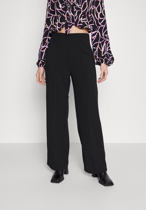 Femme portant un pantalon noir taille haute à jambes larges, des bottines noires à bout carré et un chemisier noir à manches longues avec des motifs abstraits roses et blancs.