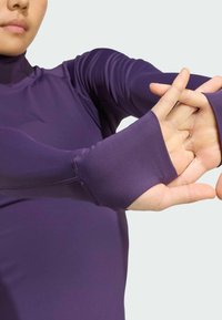 Haut de sport violet à manches longues avec orifices pour les pouces, texture lisse, design ajusté, col montant et détails de couture sur les manches.