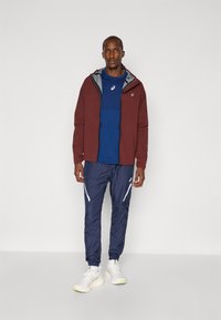 Bordeauxfarbene, leichte Jacke über einem blauen T-Shirt, kombiniert mit navyfarbenen Jogginghosen mit weißen Akzenten und weißen Sneakern. Sauberes Design, sportliche Passform.