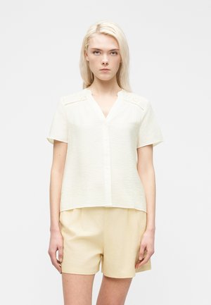 Mladá žena s rovnými blond vlasmi, oblečená v krátkemu krémovom blúzke s čipkovými detailmi a svetlobéžových kraťasoch, stojaca pred jednoduchým pozadím.