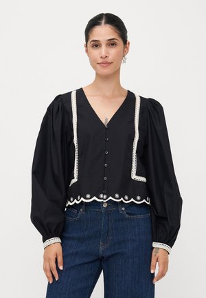 Zwarte cropped blouse met lange pofmouwen, V-hals, kanten randdetails en knoopsluiting. Decoratieve geschulpte zoom aan de onderkant.