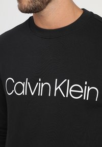 Černá bavlněná mikina s kulatým výstřihem, dlouhými rukávy a výrazným bílým logem „Calvin Klein“ přes hrudník. Hladká textura.