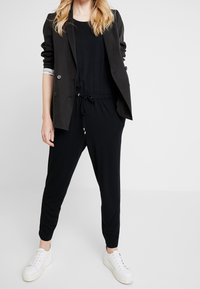 Frau in schwarzem Blazer, schwarzem Overall mit Kordelzug an der taille und weißen Plateausneakern, steht vor einem weißen Hintergrund.