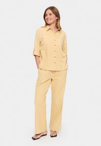 Light yellow, button-up shirt with roll-up sleeves paired with loose-fitting, matching trousers and flat sandals. Smooth, lightweight fabric.