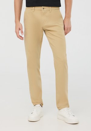 Chinos beige in cotone con design a gamba dritta, dotati di tasche frontali, chiusura con bottone e sneakers bianche.