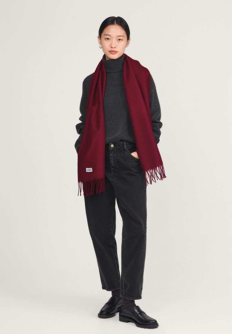 Écharpe en laine bordeaux avec des franges, drapée sur un col roulé gris foncé, accompagnée d'un jean noir taille haute et de chaussures noires.