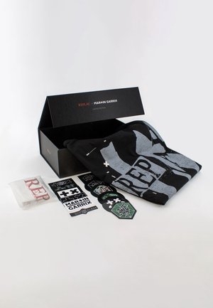 Caja negra de edición limitada etiquetada "Replay para Martin Garrix", que contiene una camiseta gráfica negra doblada, pegatinas y un pequeño objeto blanco con texto rojo.