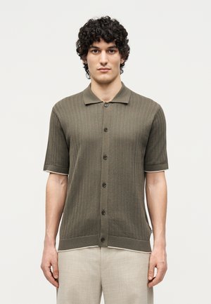 Άνδρας με σγουρά μαλλιά φοράει ελαφρύ πλεκτό πουκάμισο χρώματος ελιάς με κοντά μανίκια και ανοιχτό μπεζ παντελόνι, στέκεται μπροστά σε ένα απλό λευκό φόντο.