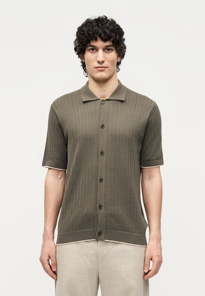 Hombre con cabello rizado lleva una camisa de punto con botones de manga corta color verde oliva y pantalones beige claro, de pie sobre un fondo blanco liso.