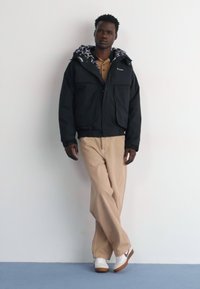 Schwarze isolierte Jacke mit einer gemusterten Kapuze, kombiniert mit beigen Hosen und weißen Turnschuhen. Das Modell steht lässig vor einem schlichten Hintergrund.