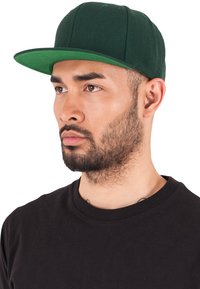 Grøn kasket med en flad skygge og en tætsiddende design, lavet af et struktureret stof, båret med en sort t-shirt, set fra siden.