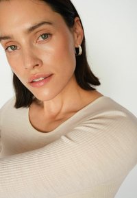 Beige gerippter Langarm-Top mit einem weitem Ausschnitt, kombiniert mit silbernen Creolen. Die Haut wirkt glatt mit einem natürlichen Glanz.