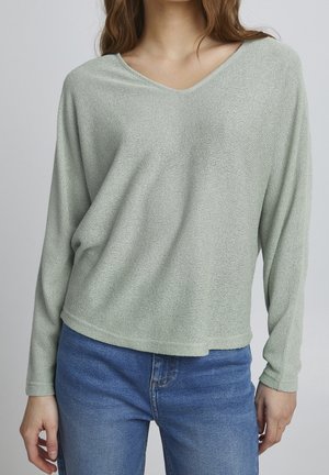 Haut vert clair à manches longues et encolure en V en tissu texturé, associé à un jean en denim bleu. Le haut a une coupe ample et un ourlet arrondi.