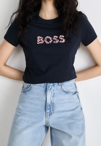 Žena oblečená v černém tričku s nápisem "BOSS" z malých růžových a bílých květin, v kombinaci s světle modrými džíny s vysokým pasem, s rukama za zády.