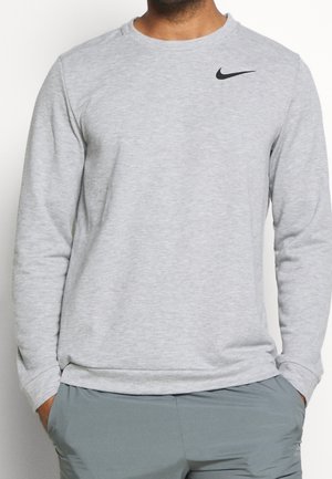 Mann trägt ein hellgraues Nike-Sweatshirt mit langen Ärmeln und schwarzem Logo, die Hände in den Taschen, kombiniert mit grauer Hose vor einfarbigem Hintergrund.
