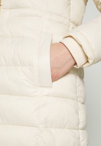 Kurtka puchowa w kolorze kremowym z poziomym, pikowanym wzorem, z boczną kieszenią i prążkowanymi mankietami, wykonana z miękkiego, błyszczącego materiału.