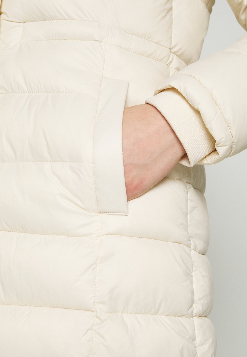 Kurtka puchowa w kolorze kremowym z poziomym, pikowanym wzorem, z boczną kieszenią i prążkowanymi mankietami, wykonana z miękkiego, błyszczącego materiału.