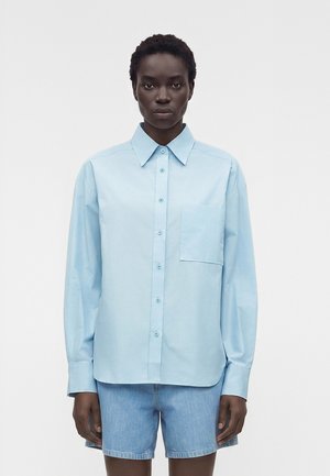 Μοντέλο με κοντά μαλλιά φοράει ανοιχτά γαλάζιο πουκάμισο με κουμπιά και τσέπη στο στήθος και ανοιχτά γαλάζια τζιν σορτς σε λευκό φόντο.