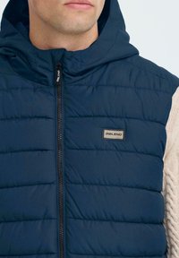 Navy modrý vypolstrovaný vest, který má zipové zapínání, horizontálně prošívané vzory a logovou nášivku na hrudi, kombinovaný s texturovaným svetrem.