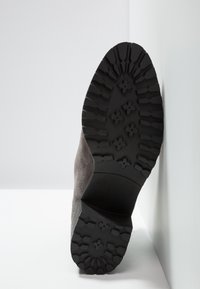 Semelle d'une botte en suède gris montrant une semelle extérieure épaisse en caoutchouc noir avec des rainures à motifs et un talon bloc sur fond blanc.