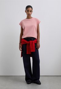 Różowy t-shirt z krótkim rękawem, w połączeniu z ciemnymi, szerokimi spodniami dżinsowymi oraz czerwonym, dzianinowym swetrem zawiązanym w talii, z detalami teksturowanej tkaniny.