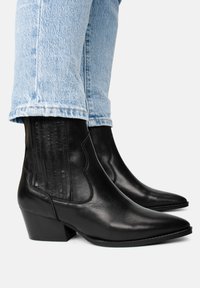 Botas de cuero negras hasta el tobillo con punta afilada, tacón grueso, paneles laterales decorativos y un acabado suave. Llevadas con jeans de color azul claro.
