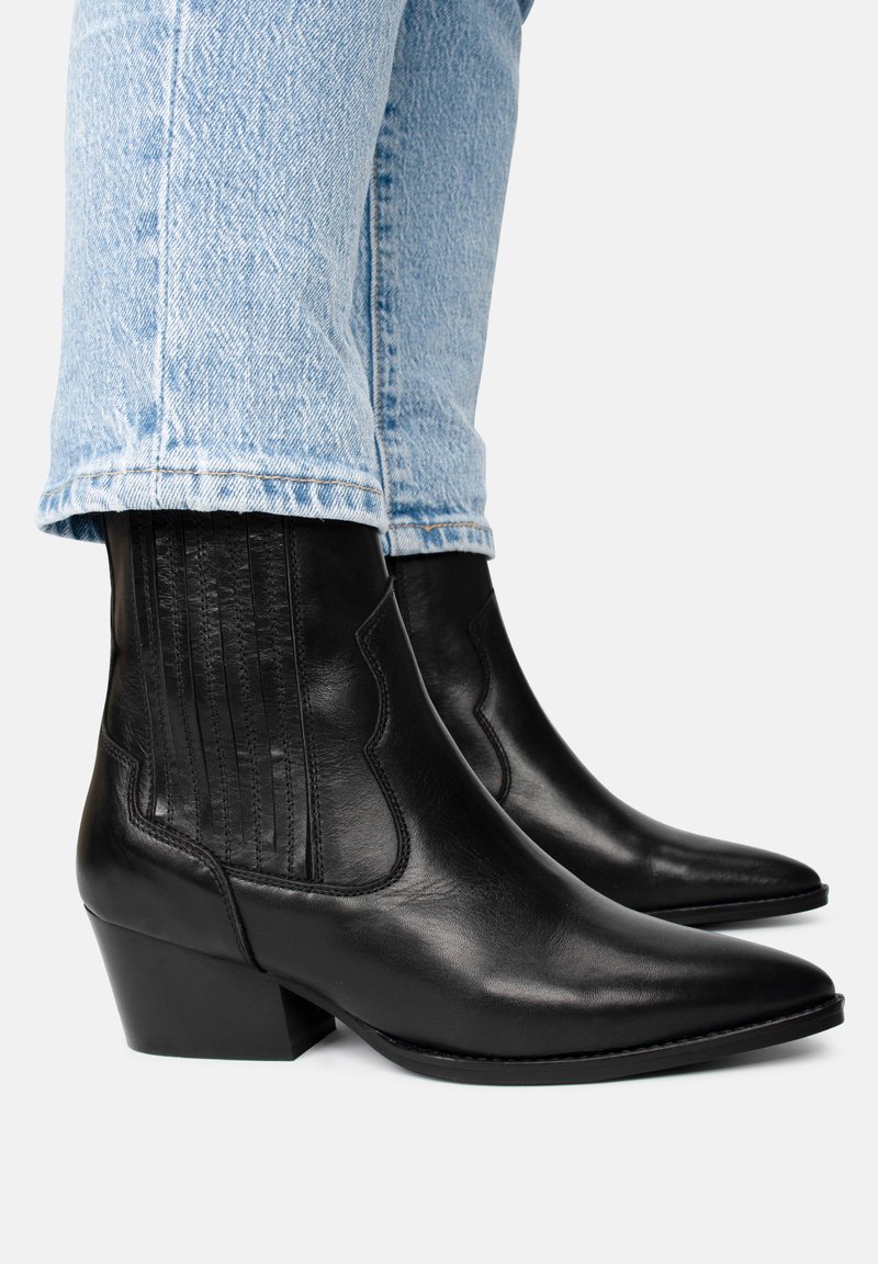 Botas de cuero negras hasta el tobillo con punta afilada, tacón grueso, paneles laterales decorativos y un acabado suave. Llevadas con jeans de color azul claro.