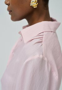 Camisa de color rosa claro con un collar estructurado, sutiles rayas verticales y detalles fruncidos en los hombros. Acompañada de un arete abstracto dorado.