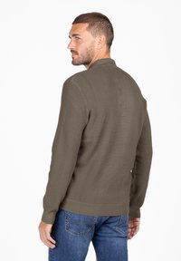 Olivegrüner, strukturierter Pullover mit einem gerippten Kragen und Bündchen, kombiniert mit blauen Jeans. Das Design zeichnet sich durch einen klaren, minimalistischen Look aus.