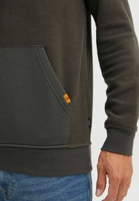 Sudadera de forro polar en verde oliva oscuro con un bolsillo gris en contraste, puños acanalados y una etiqueta de acento naranja en el borde del bolsillo.