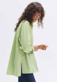 Light green long-sleeve shirt with a relaxed fit, featuring button cuffs, side slits, and smooth fabric texture. Worn with blue jeans.