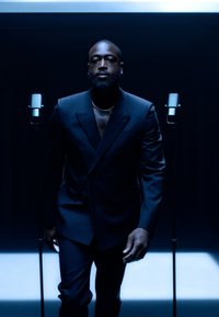 Svart skräddarsydd kostym med slagkrage, som bärs över en bar bröstkorg. Ett silverkedja hängande runt halsen och två mikrofoner i en mörk miljö.