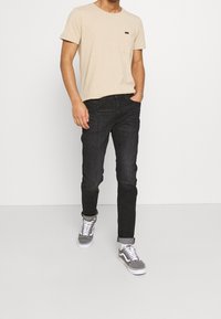 Homme portant un t-shirt beige à manches courtes, un jean noir avec ourlets, et des baskets basses grises, se tenant contre un fond blanc.