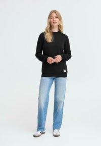 Felpa nera con scollo rotondo e polsini a coste, abbinata a jeans azzurri chiari e sneakers bianche con dettagli grigi. Presentazione di un outfit casual.
