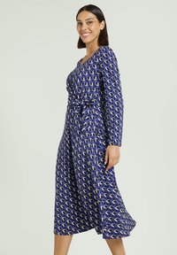 Niebieska sukienka z długim rękawem o geometrycznym wzorze w jasnoniebieskim, białym i fioletowym kolorze. Posiada pasek do wiązania i dekolt w kształcie litery V.